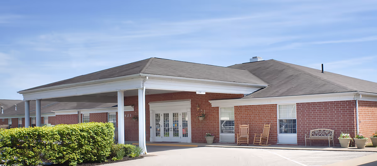 Exterior view of a single-story brick building with a covered entrance supported by white columns. There are two rocking chairs and a bench near the entrance, along with several potted plants. The sky is clear and blue.
