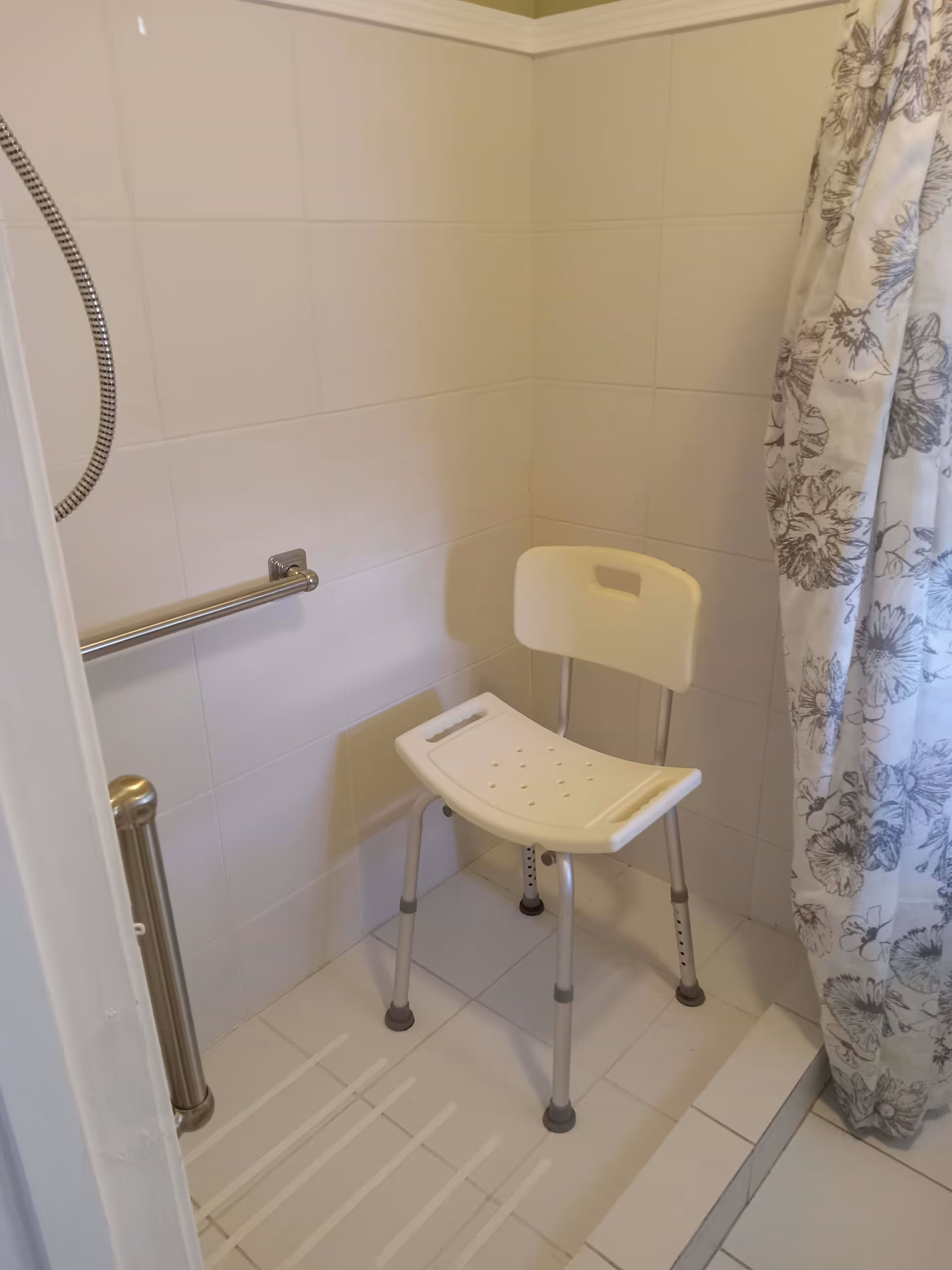 A small shower area with white tiled walls and floor, featuring a white shower chair with a backrest and adjustable legs, a silver grab bar on the wall, and a floral-patterned shower curtain partially visible on the right side.