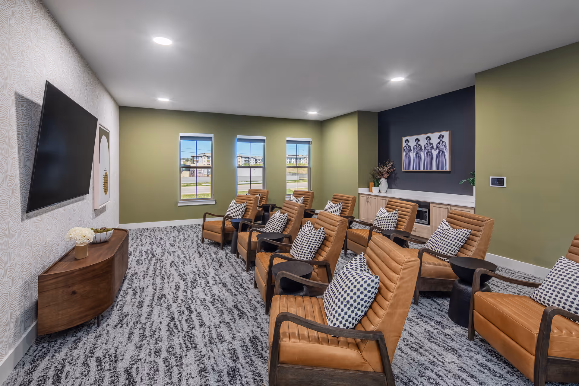 A well-lit media lounge with rows of tan leather chairs and patterned pillows facing a wall-mounted TV.