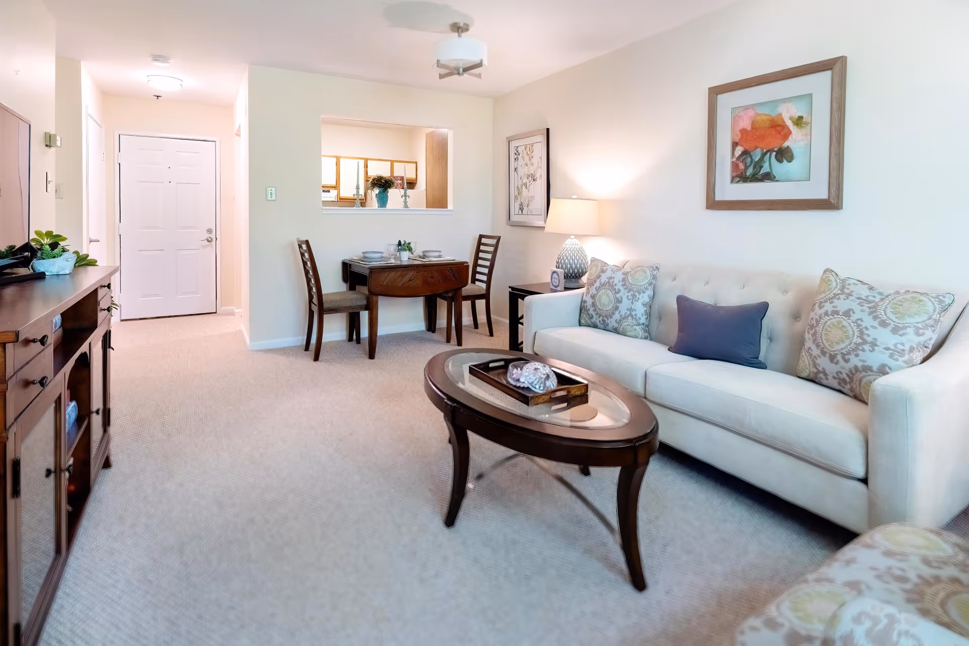 A bright and cozy living room with a beige sofa adorned with patterned and solid pillows, a wooden coffee table with decorative items, a side table with a lamp, and framed artwork on the walls. Adjacent to the living room is a small dining area with a wooden table and two chairs, and a pass-through window to the kitchen. The entrance door is visible in the background.