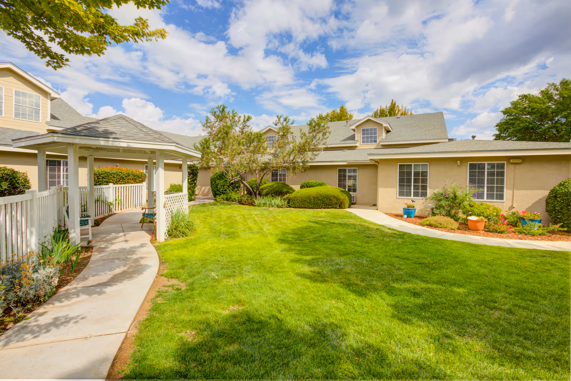A bright outdoor garden area at Avista Senior Living Albuquerque featuring a well-maintained green lawn, a paved walkway leading to a white gazebo, various shrubs and potted plants, and beige buildings with multiple windows under a partly cloudy sky.