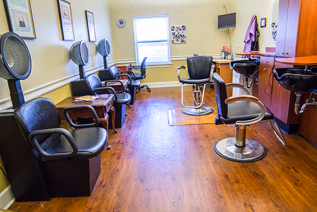 Interior of a salon area in a senior living facility with multiple black salon chairs, hair drying stations along the left wall, and a counter with mirrors and sinks on the right. The room has wooden flooring, a window at the far end, and framed pictures on the walls.