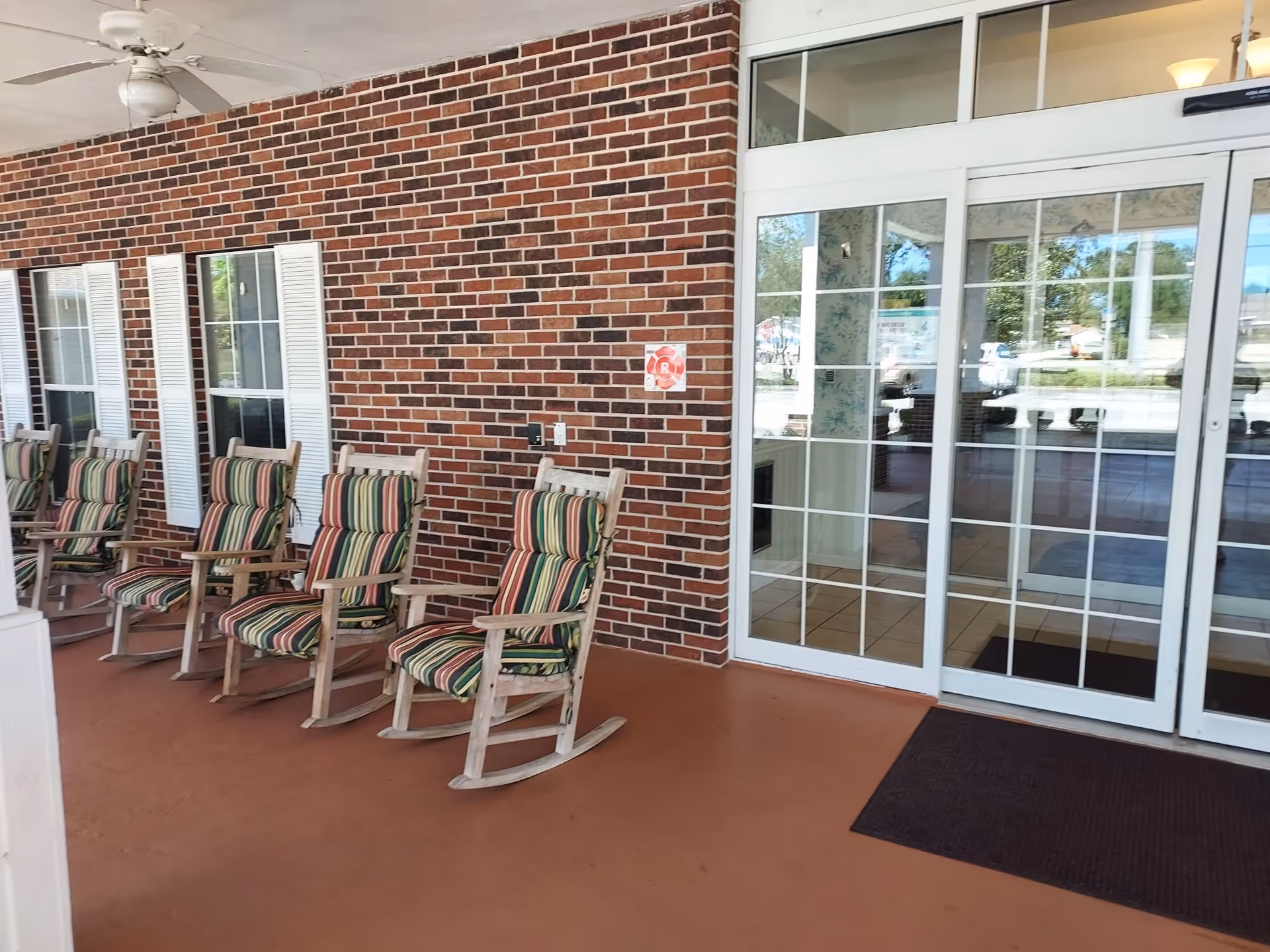 Covered entrance porch with a row of striped-cushioned wooden rocking chairs along a brick wall next to glass double doors.