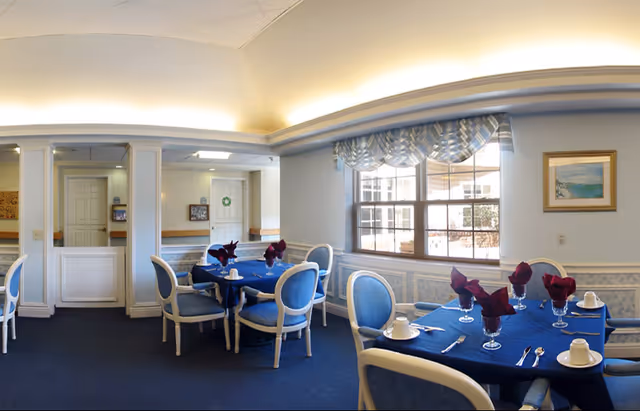 Dining room with round tables covered in blue tablecloths, blue upholstered chairs, place settings with folded napkins, and a large window with a valance.