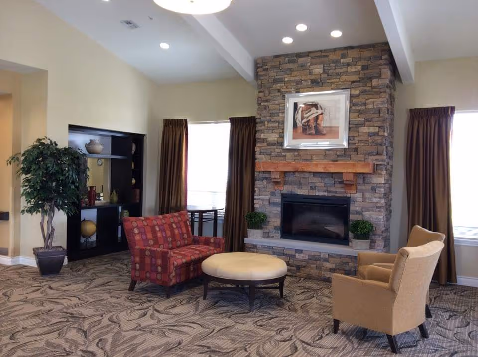 A cozy living room area with a stone fireplace in the center, flanked by two windows with brown curtains. In front of the fireplace is a round beige ottoman, a red patterned loveseat, and a beige armchair. To the left, there is a tall potted plant and a black shelving unit with decorative items. The room has a patterned carpet and recessed ceiling lights.