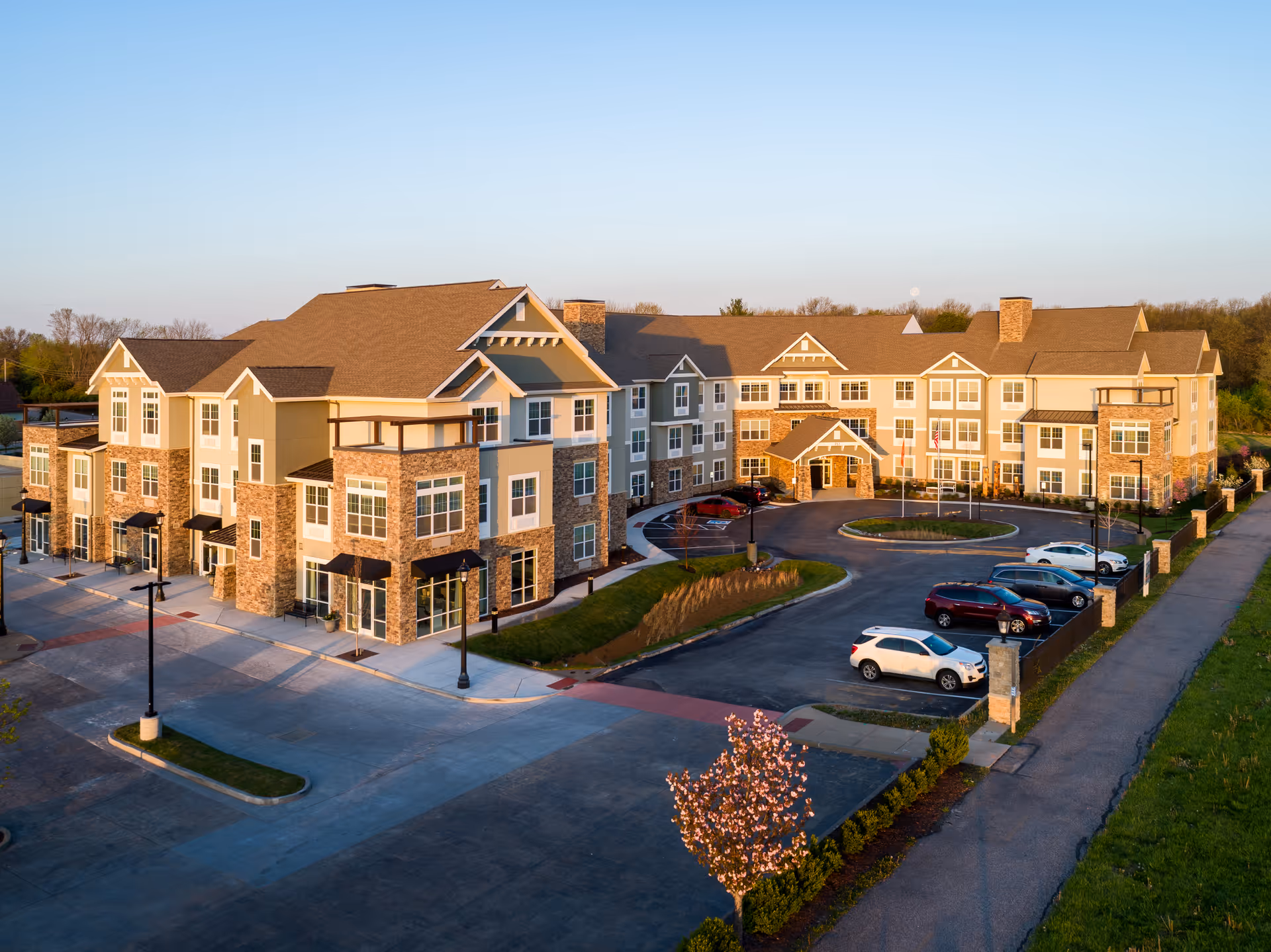 A large, multi-story senior living facility building with beige and stone exterior walls, multiple windows, and a pitched roof. The building is surrounded by a parking lot with several cars parked, a circular driveway, and landscaped greenery including a blooming tree in the foreground. The sky is clear and the scene is lit by warm sunlight.