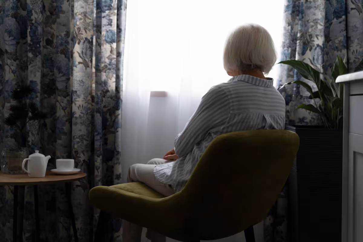 An elderly person with white hair sitting on a green chair facing a window with sheer white curtains and floral-patterned drapes. A small round table with a white teapot and cup is beside them, and a green plant is visible in the background.