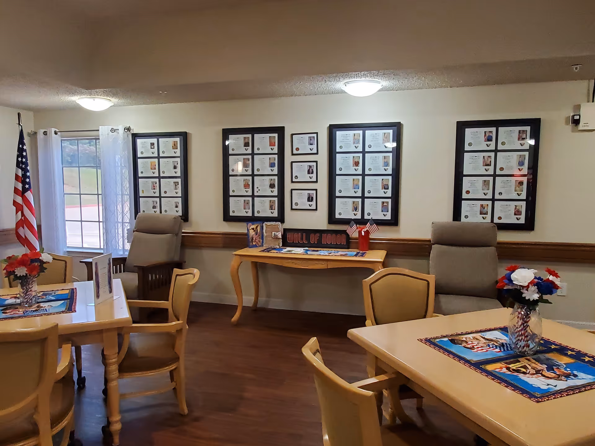 Interior room with tables and chairs arranged for seating, decorated with patriotic-themed placemats and flower vases. Two comfortable armchairs are placed against the wall, which features a 'Wall of Honor' display with framed photos and certificates. An American flag stands near a window with sheer white curtains, and the room has warm lighting and wood flooring.