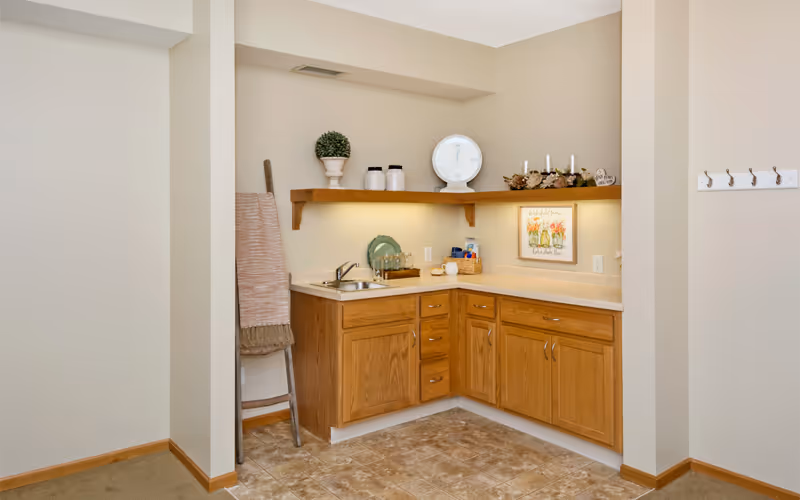 A small kitchenette area with wooden cabinets and a countertop featuring a sink, a dish rack with plates, and various decorative items including a potted plant, jars, a scale, and a framed floral picture. There is a wooden shelf above the countertop with candles and other decor. A ladder with a folded blanket leans against the wall to the left, and a wall-mounted coat rack with hooks is visible on the right.