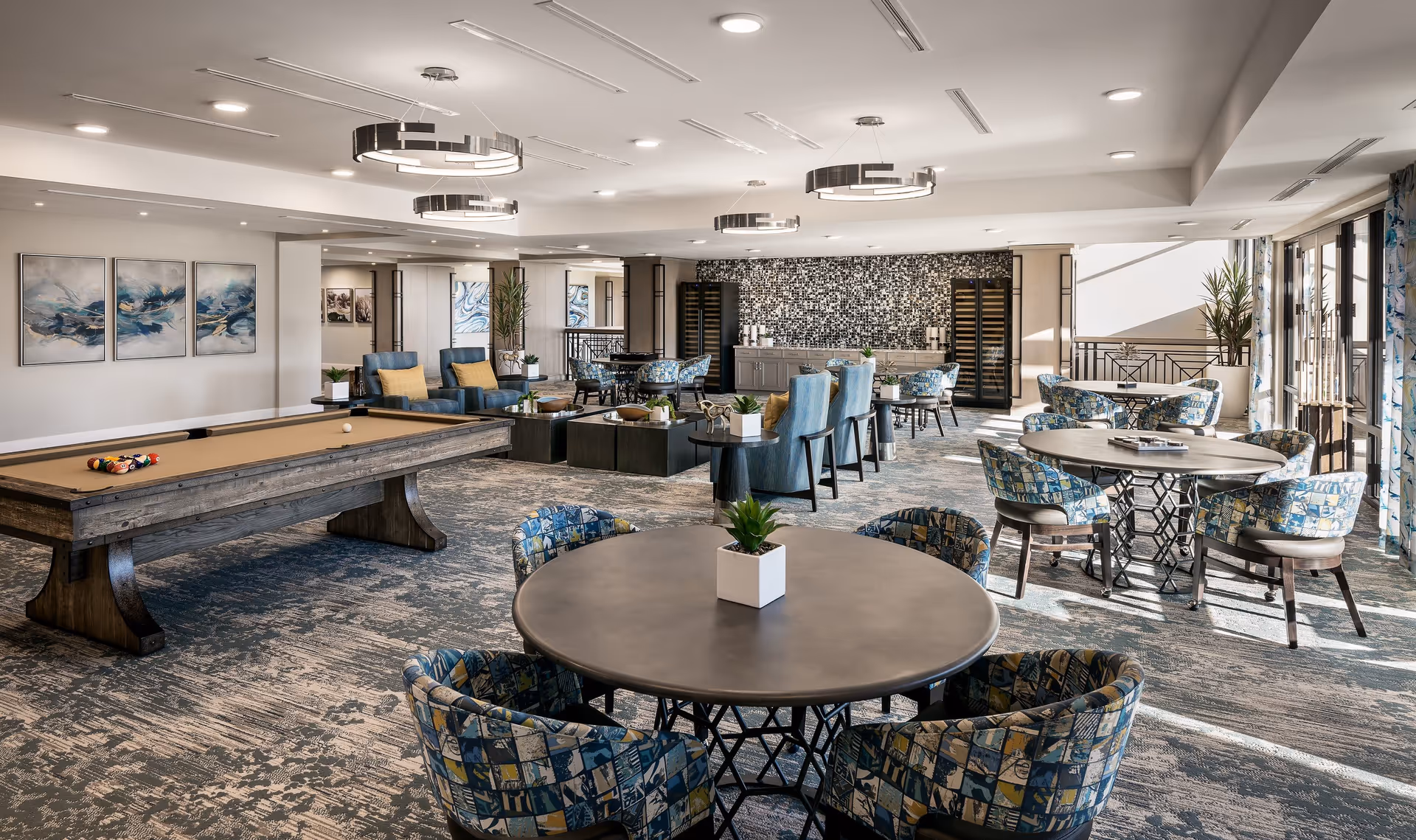 Spacious, well-lit communal lounge with a pool table, round tables and patterned upholstered chairs, lounge seating, and a beverage/service counter.