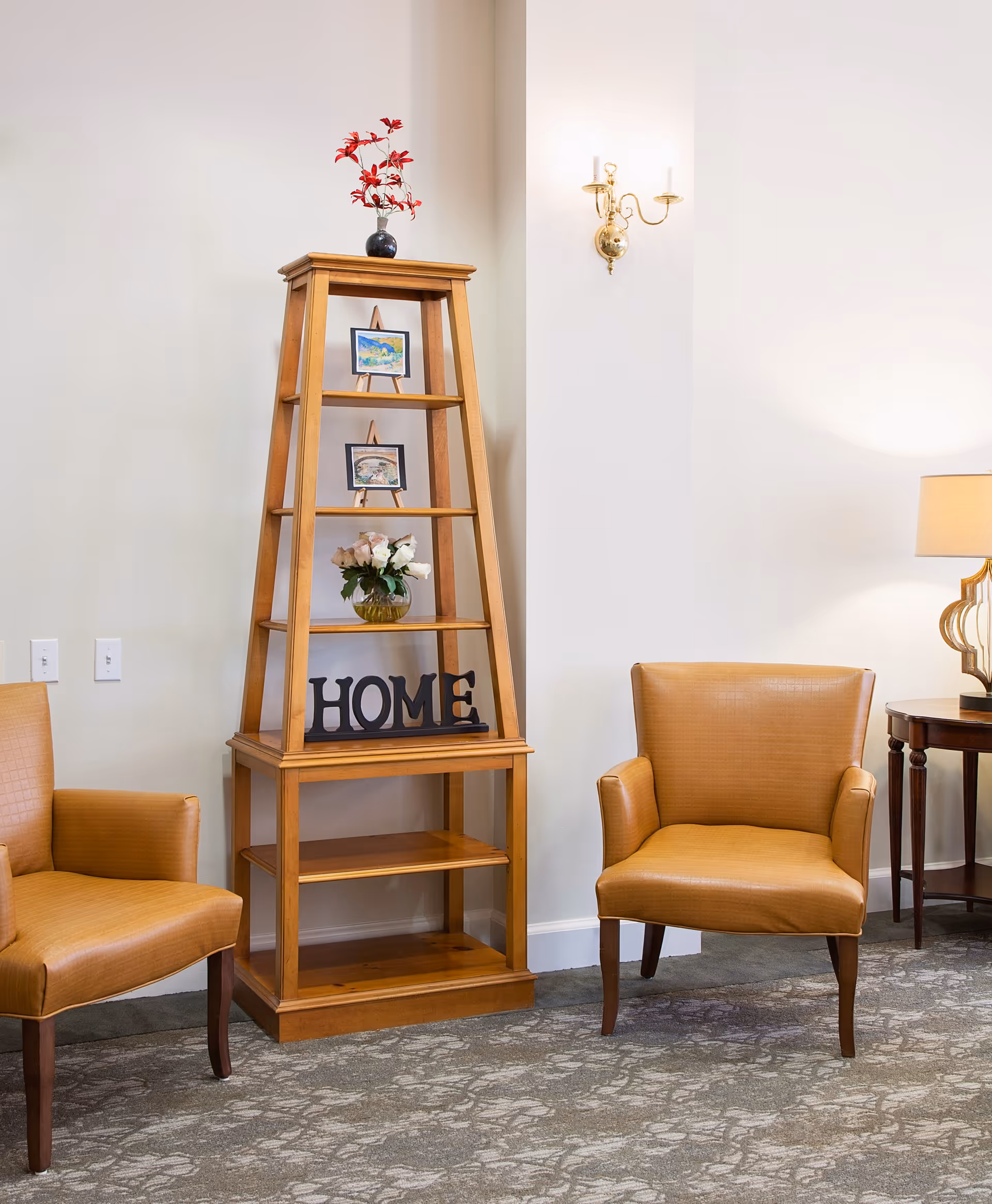 A cozy corner of a living room featuring two tan leather armchairs on a patterned carpet. Between the chairs is a wooden shelving unit with decorative items including a vase with flowers, two small framed pictures, and a decorative 'HOME' sign. A wall-mounted brass sconce and a table lamp on a side table provide warm lighting.