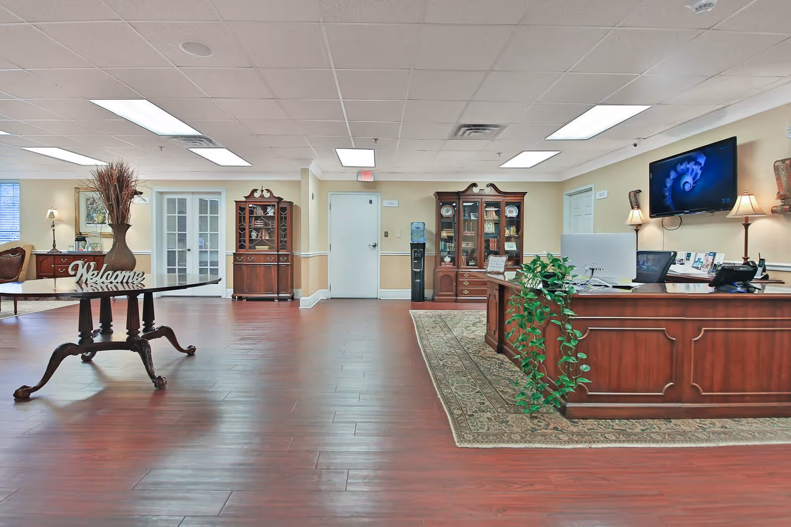 A spacious reception area with polished wooden floors and beige walls. There is a large wooden reception desk with a computer and a green plant on it, positioned on a patterned rug. Behind the desk is a wall-mounted TV and two table lamps. To the left, there is a wooden table with a decorative vase and a 'Welcome' sign. Two wooden display cabinets with glass doors are against the far wall, and a water dispenser is placed between them. The ceiling has recessed lighting panels.