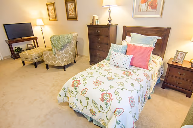 Well-lit bedroom with a twin bed dressed in a floral bedspread, nightstands and dresser, plus an armchair and TV.