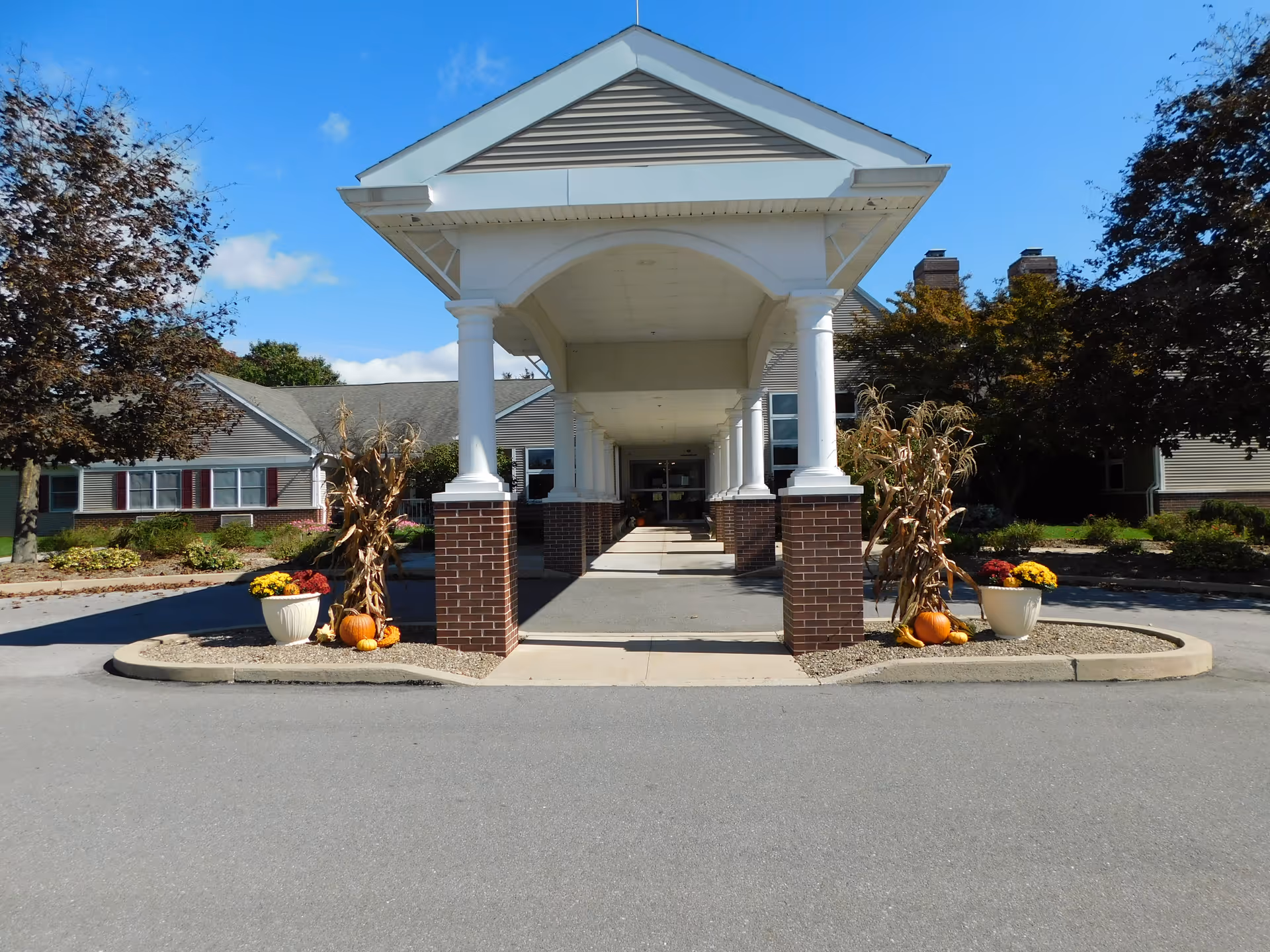 Covered entrance canopy with white columns and brick bases leading to the front door of a senior living facility, flanked by pumpkins and planters.