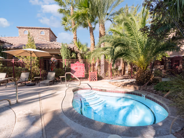 Outdoor hot tub or spa surrounded by palm trees and lounge chairs with umbrellas, adjacent to a building with a tiled roof under a blue sky with some clouds.