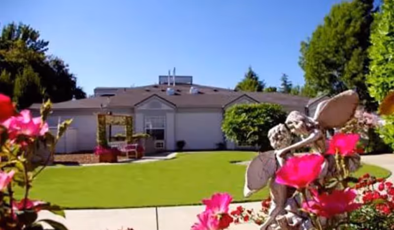Outdoor garden area with a well-maintained lawn, colorful flowers in the foreground, a statue of two fairies, and a single-story building in the background under a clear blue sky.