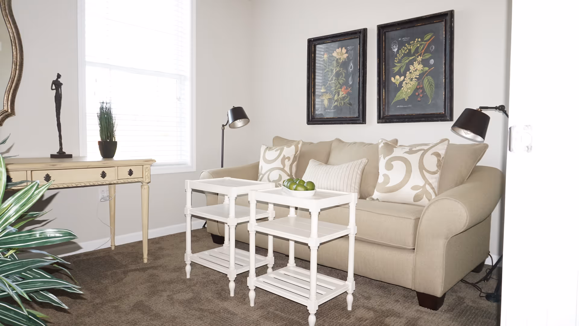 Bright living room with a beige sofa, two white nesting side tables, framed botanical prints, and a console table by a window.