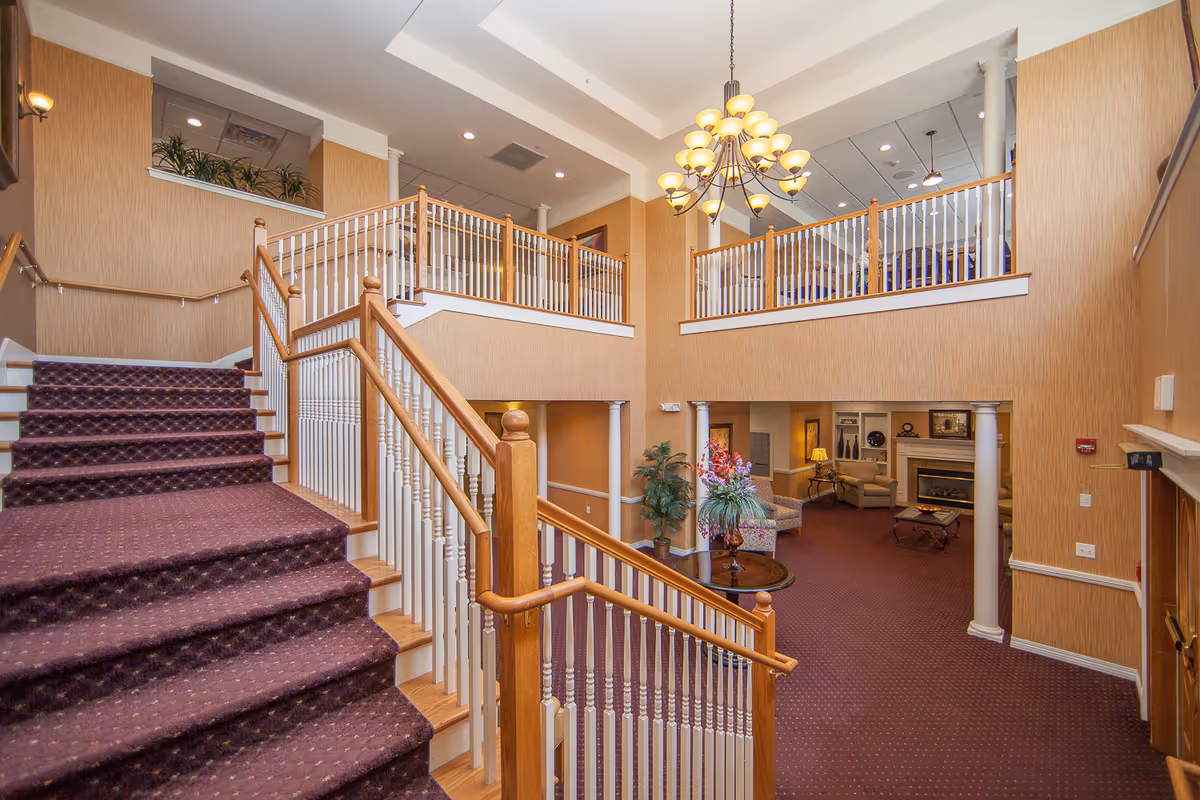 Interior view of a senior living facility featuring a carpeted staircase with wooden handrails leading to an upper level. The area has beige walls, a chandelier hanging from the ceiling, and a sitting area with chairs and a fireplace visible in the background.