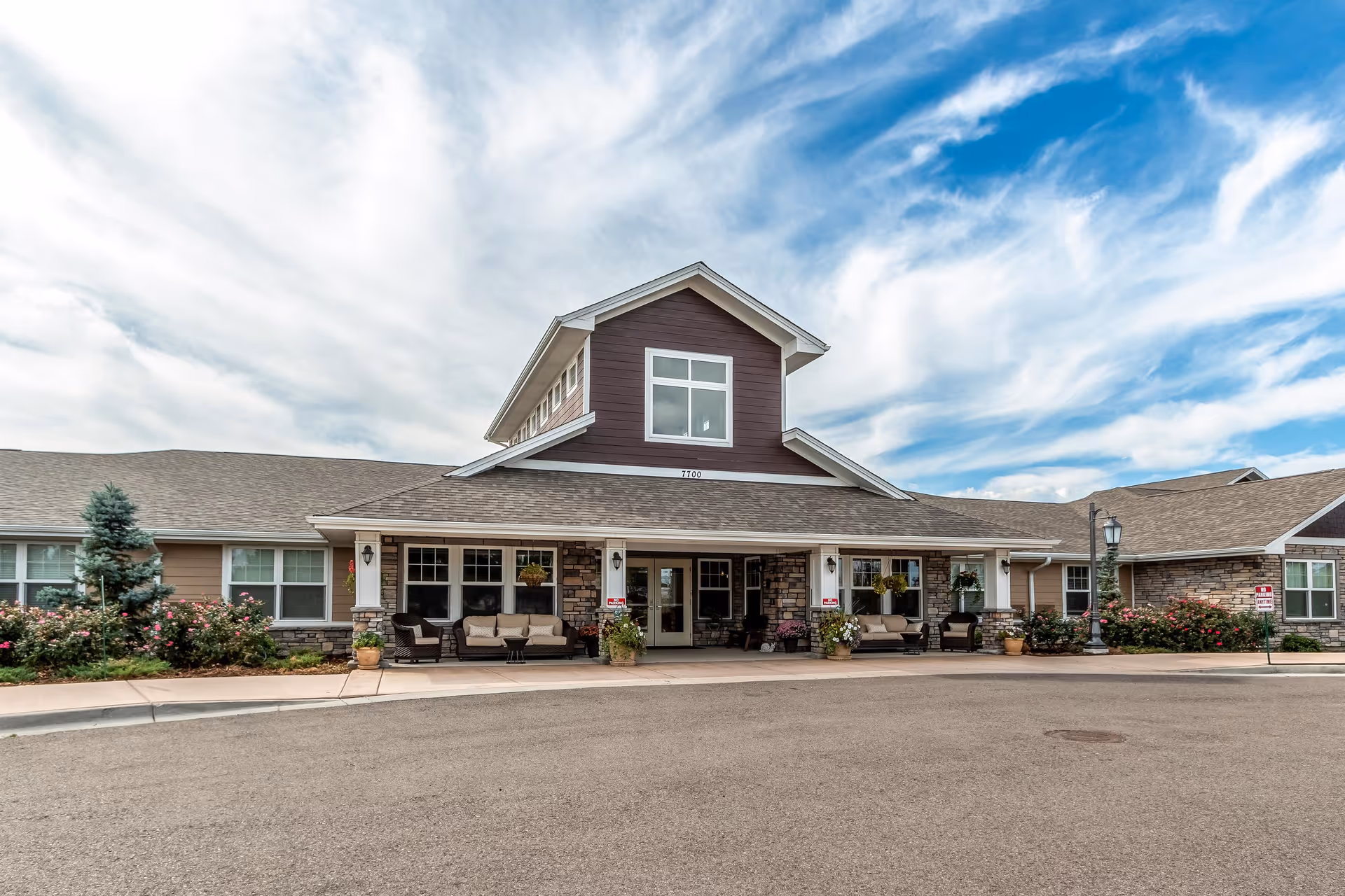 Front exterior view of a single-story senior living facility building with a covered entrance, outdoor seating, flower pots, and landscaping under a partly cloudy blue sky.
