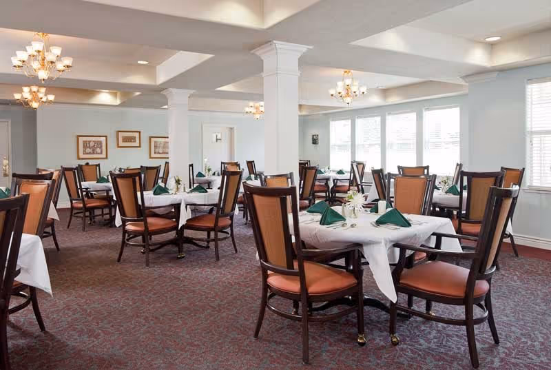 A spacious dining room with multiple round tables covered with white tablecloths, each set with green folded napkins and small floral centerpieces. The room features wooden chairs with orange cushions, carpeted floors, white walls, large windows letting in natural light, and elegant chandeliers hanging from the ceiling.