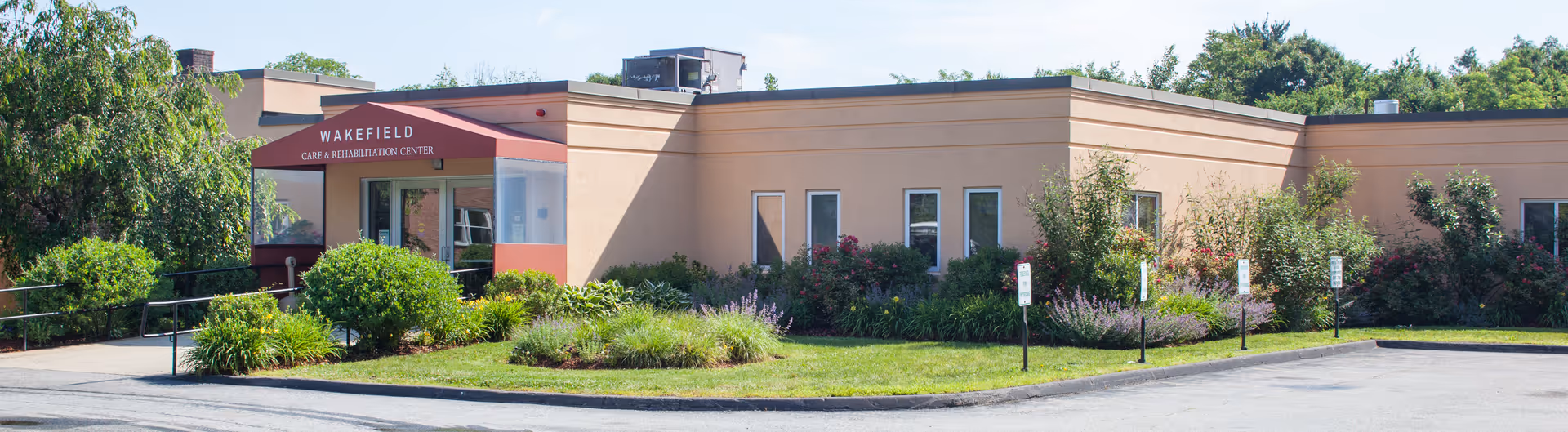 Front entrance of a single-story care and rehabilitation center with a red awning reading 'WAKEFIELD,' landscaped bushes, and a parking area.