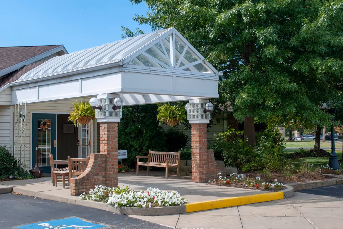 Entrance area of a facility with a covered porch supported by brick columns, hanging plants, wooden benches, and a small flower bed. There is a building with a door and windows in the background, surrounded by trees and greenery.