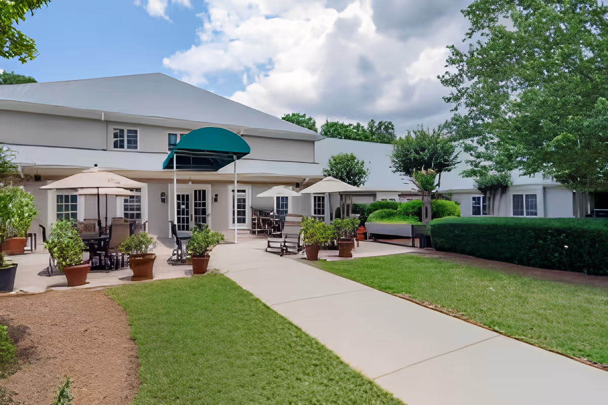Outdoor patio area at Charter Senior Living of Buford with tables, chairs, and umbrellas surrounded by potted plants and greenery, leading to a white building under a partly cloudy sky.