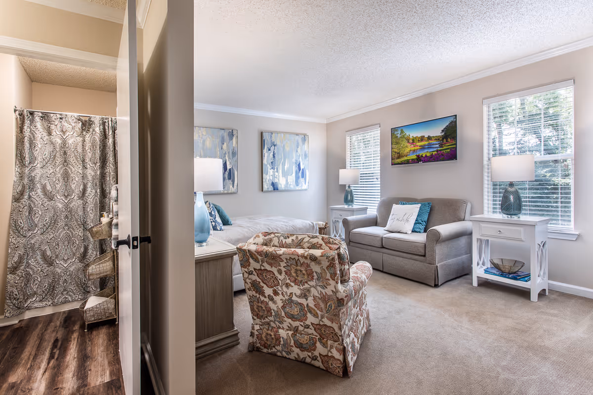 A cozy living space with a gray loveseat, a floral patterned armchair, two white side tables with lamps, and two abstract paintings on the wall. There are two windows with blinds letting in natural light. A bathroom with a patterned shower curtain is visible through an open door.