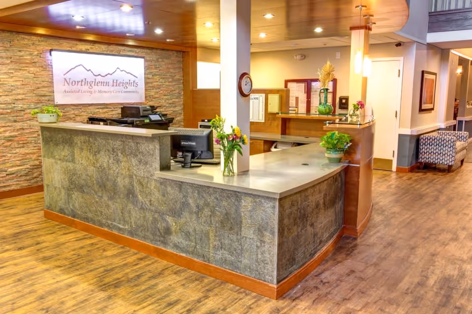 Reception area of Northglenn Heights Assisted Living & Memory Care Community featuring a stone and wood front desk with computer monitors, flower vases, and a clock on a white pillar. The background includes a stone accent wall with a lit sign displaying the facility name, wood flooring, and a seating area with patterned chairs.