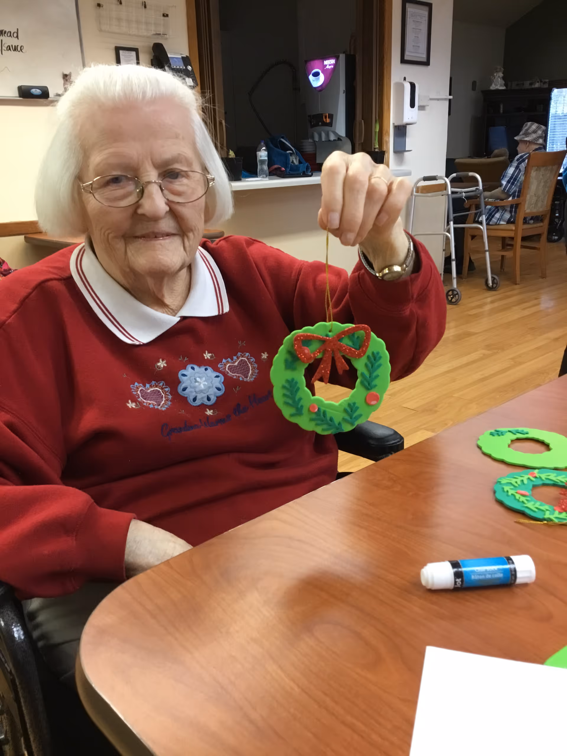 An elderly woman with white hair and glasses is sitting at a wooden table in a common area. She is wearing a red sweater with decorative embroidery and holding up a green craft wreath decorated with a red bow and holly. On the table are additional green wreath crafts and a glue stick. In the background, another elderly person is seated near a walker, and there is a coffee machine and some furniture visible.
