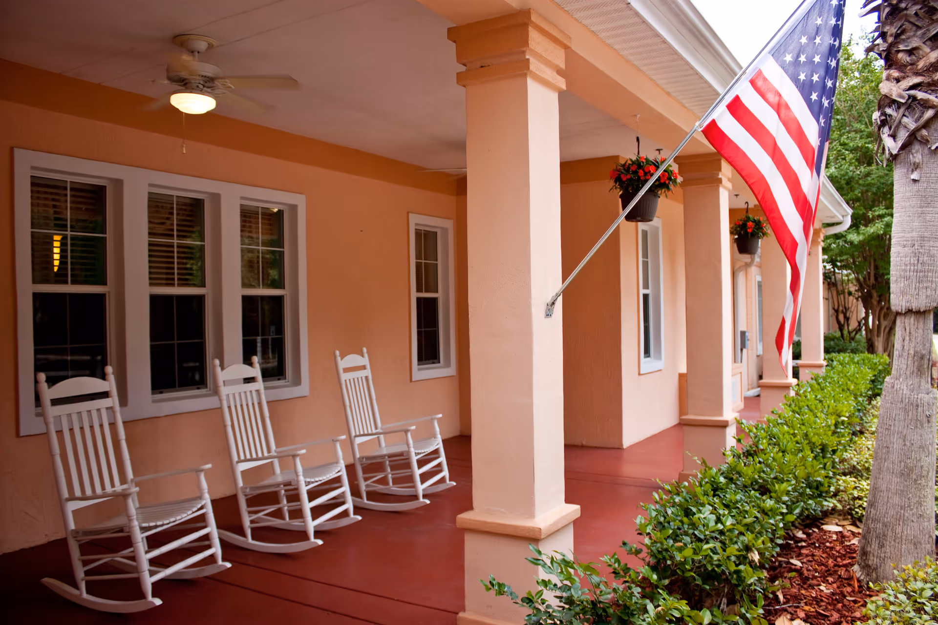 A covered porch area with three white rocking chairs lined up against a peach-colored wall with windows. There is a ceiling fan with a light above the chairs. An American flag is mounted on a pole attached to one of the porch columns. Green shrubs and a palm tree are visible along the edge of the porch.