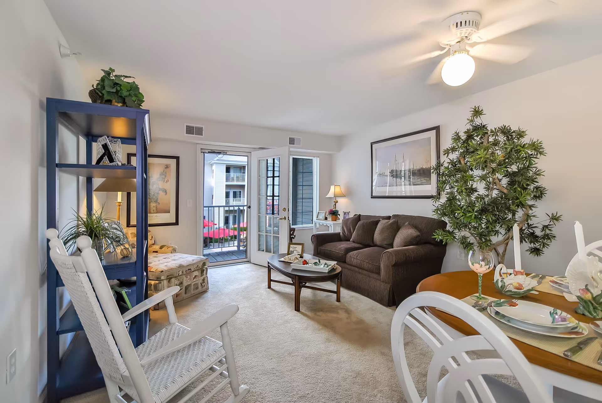 A bright and cozy living room in a retirement community apartment featuring a brown sofa, a wooden coffee table, a white rocking chair, a blue shelving unit with plants and decor, a small dining table set with plates and glasses, and a large potted plant. French doors open to a balcony with a view of neighboring buildings and red umbrellas.