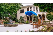 Outdoor garden area of a senior living facility with a stone fountain, manicured bushes, trees, flowers, a wooden bench, and a building with arched windows and a blue awning in the background.