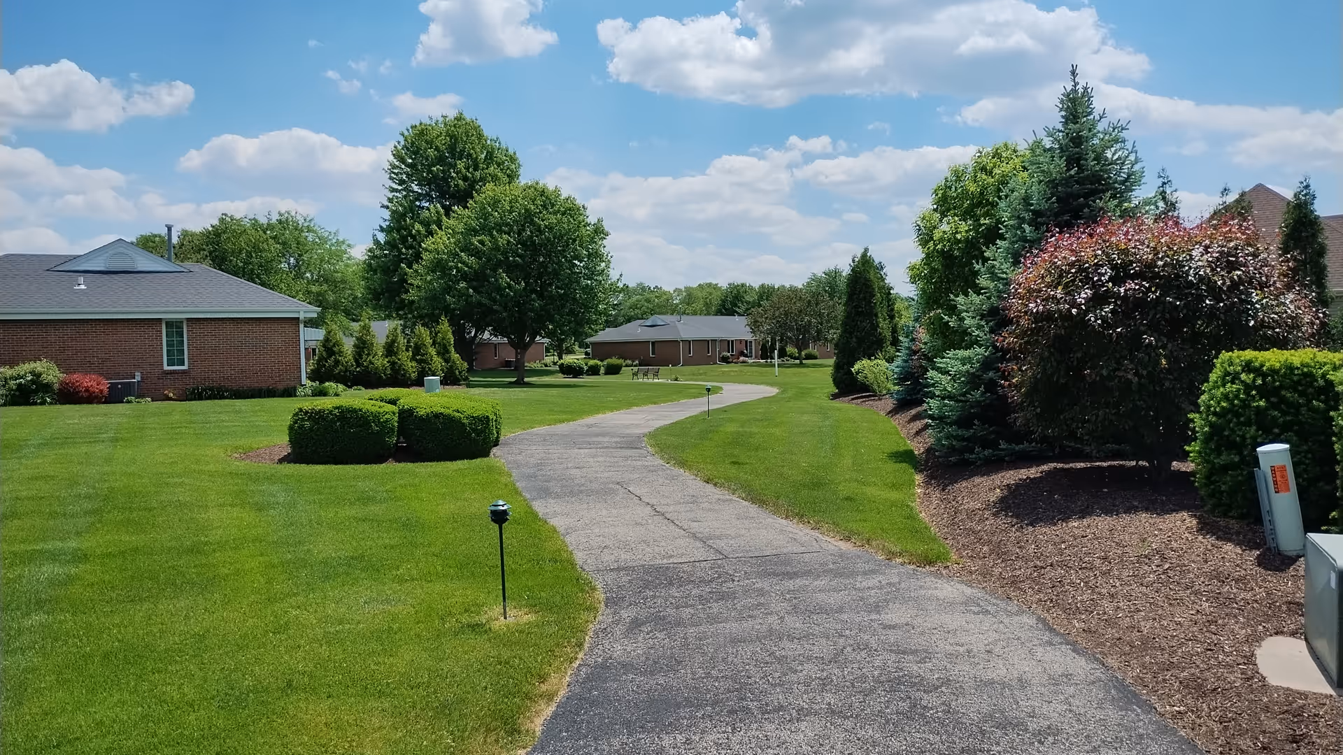 A winding paved pathway through a well-maintained green lawn with various trees and shrubs on both sides under a partly cloudy blue sky. Single-story brick buildings are visible in the background.