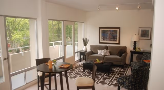 A cozy living room with a gray sofa, patterned armchair, black ottoman, and a small round dining table with chairs near large windows and a glass door leading to a balcony. The room is decorated with framed artwork, a table lamp, and a patterned rug.