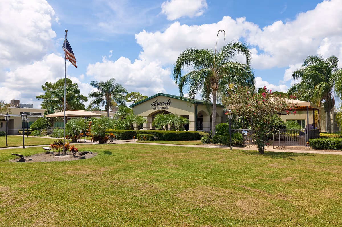 Exterior view of Avante at Orlando senior living facility with a well-maintained lawn, palm trees, an American flag on a flagpole, and a building entrance under a partly cloudy sky.