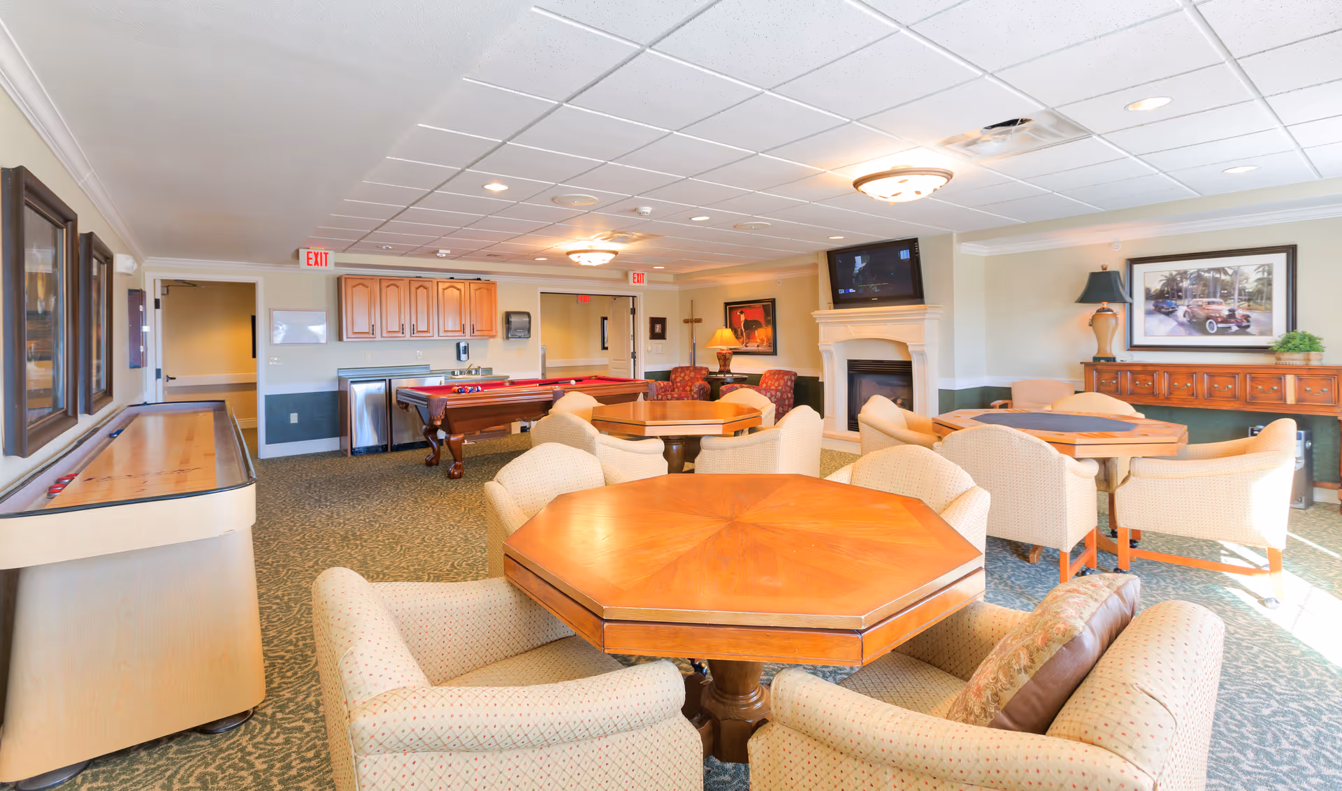 Bright communal lounge with multiple octagonal game tables and upholstered chairs, a fireplace and TV, plus a pool table and shuffleboard.