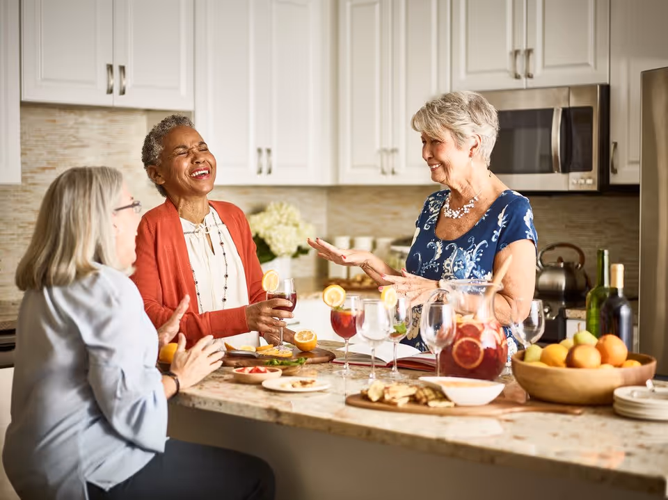 Three elderly women enjoying a lively conversation and drinks in a modern kitchen with white cabinets and a granite countertop. The countertop has a pitcher of fruit-infused beverage, glasses with lemon slices, a bowl of oranges, and plates with snacks.
