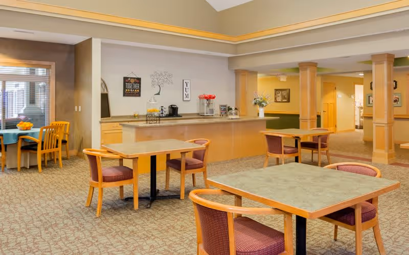 A spacious dining area in an assisted living facility with several square tables and wooden chairs with purple cushions. There is a counter with a coffee maker, a popcorn machine, and decorative wall art. The room has beige walls, carpeted floors, and large windows letting in natural light.