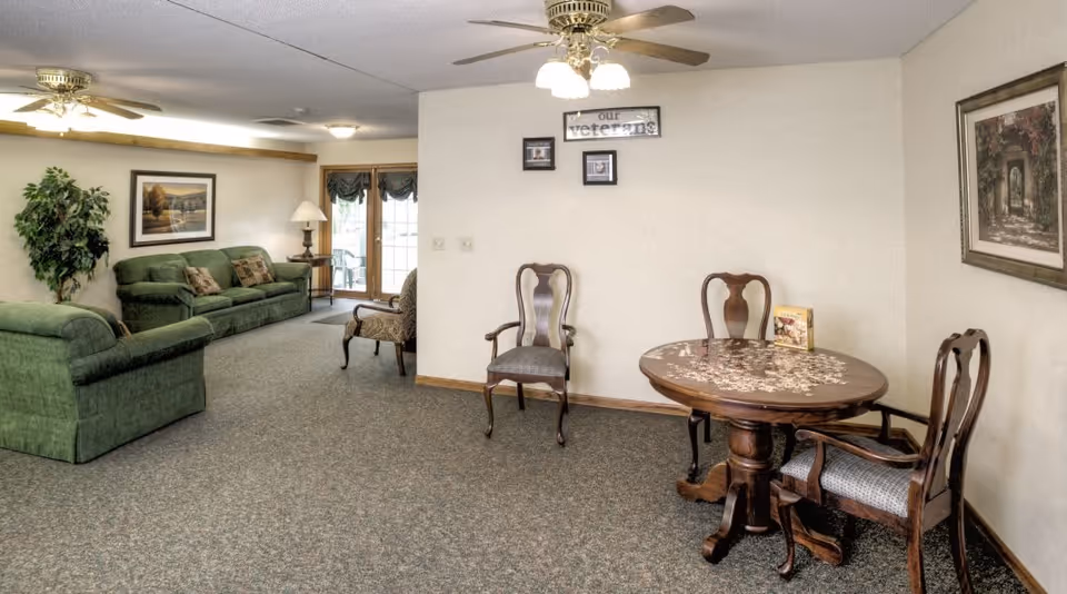 A senior living facility common area with green upholstered sofas, a wooden round table with three chairs, framed artwork on the walls, a ceiling fan with lights, and a potted plant near a set of glass doors leading outside.