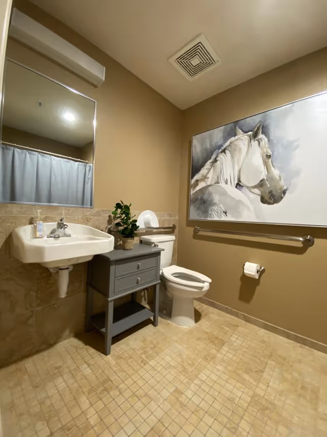 A clean bathroom with beige tiled floor and walls. It features a white wall-mounted sink with a soap dispenser, a gray cabinet with drawers, a white toilet with a roll of toilet paper on a holder, and a large painting of a white horse on the wall. There is also a mirror above the sink and a ventilation vent on the ceiling.