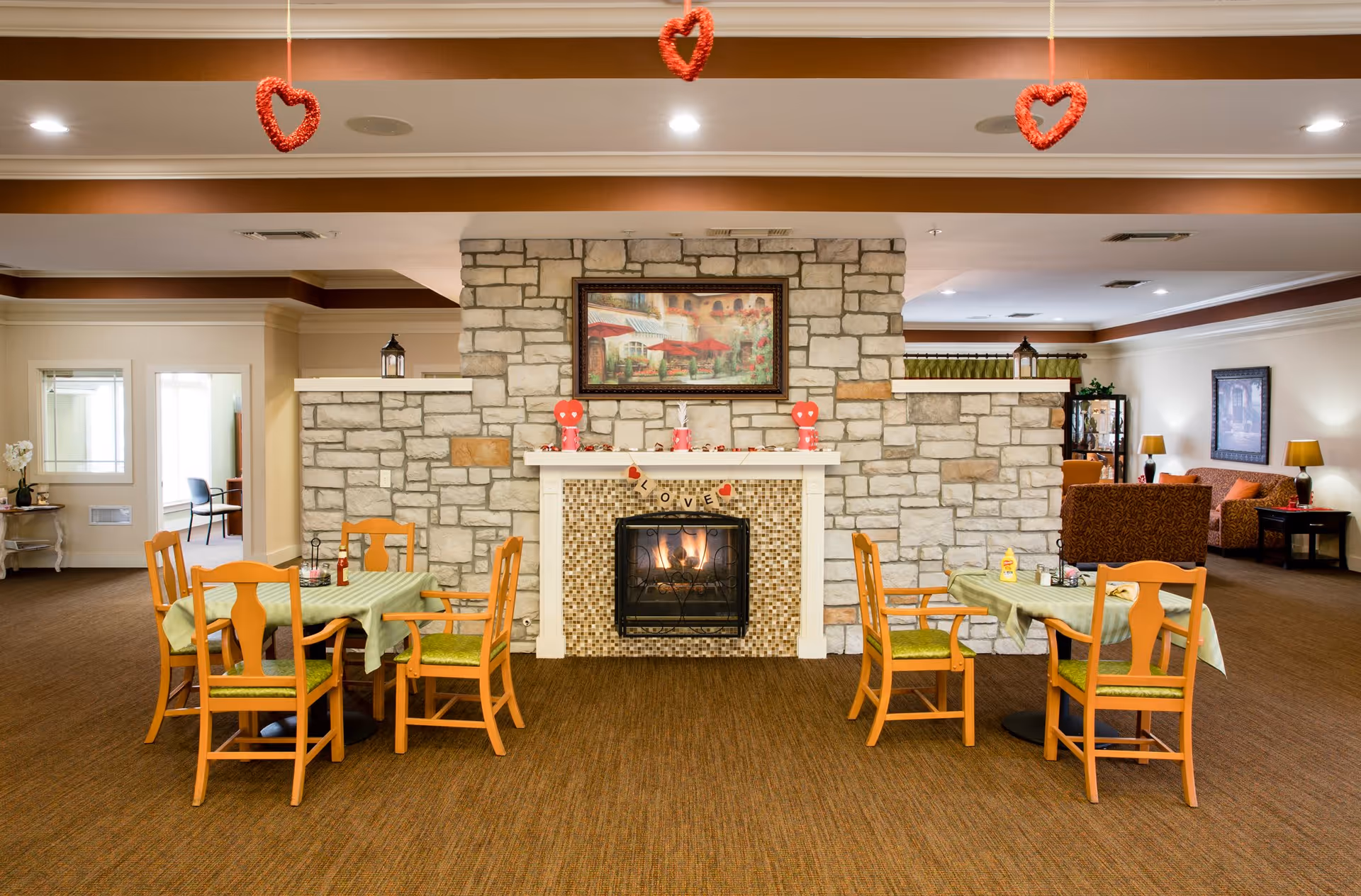 A cozy common area in an assisted living facility featuring a stone fireplace with a fire burning inside. Two dining tables with green tablecloths and wooden chairs are positioned in front of the fireplace. Red heart decorations hang from the ceiling. In the background, there is a seating area with sofas and lamps, and a framed painting above the fireplace.
