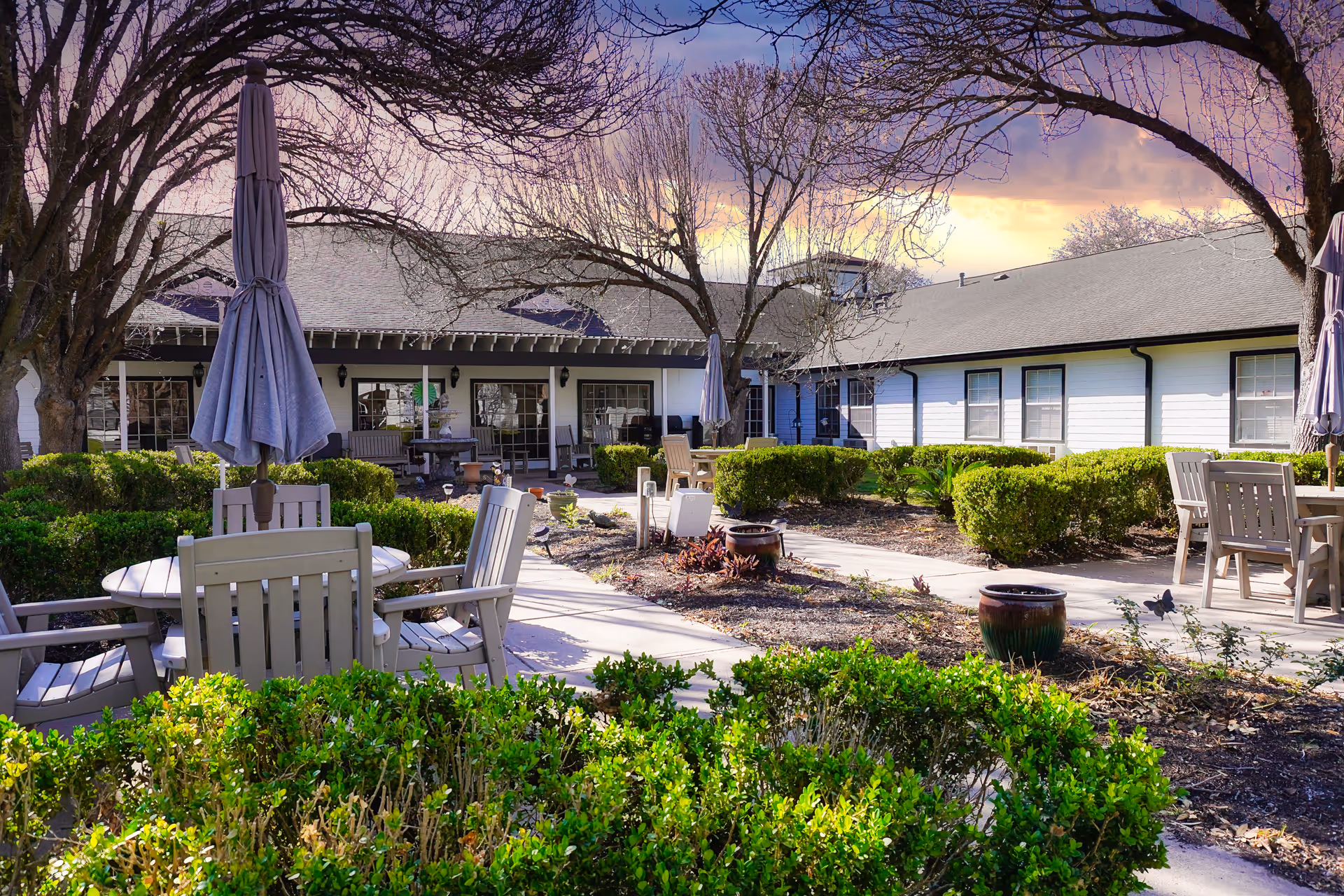 Outdoor courtyard with round tables, chairs and closed umbrellas amid landscaped shrubs in front of a single-story assisted living building.