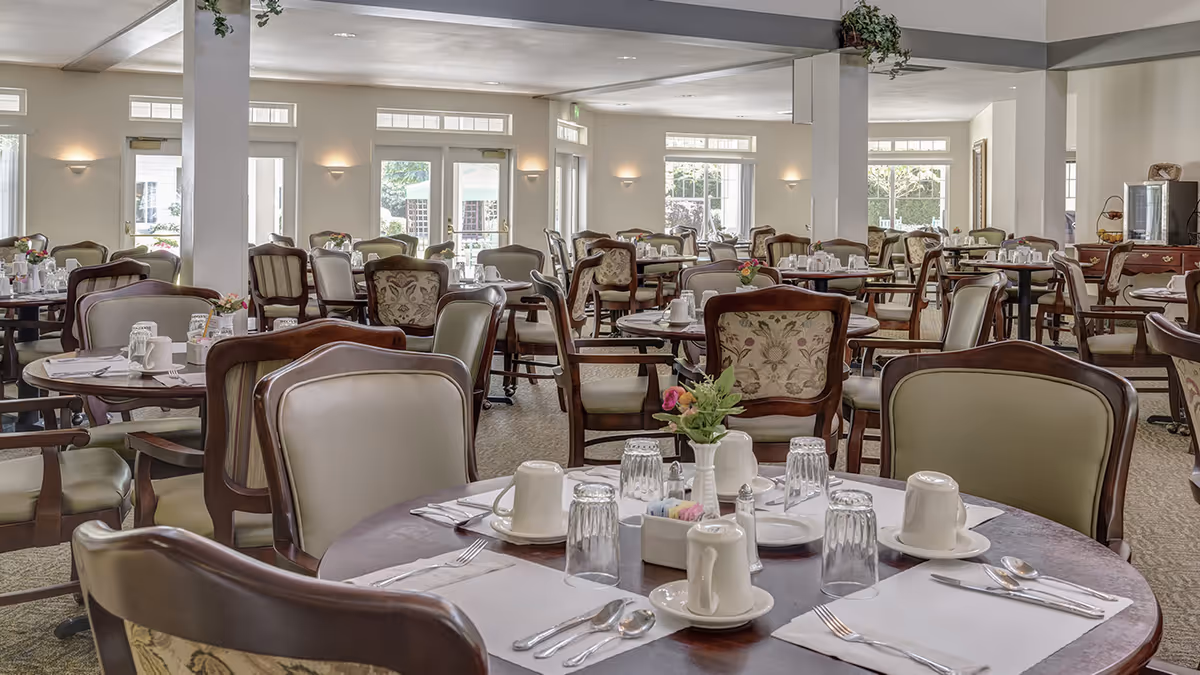 A spacious dining room with multiple round tables set with white placemats, cups, glasses, silverware, and small flower vases. The room has large windows and glass doors allowing natural light to fill the space. The chairs have wooden frames with cushioned seats and backs, some with floral upholstery. The walls are light-colored, and there are wall-mounted lights providing additional illumination.