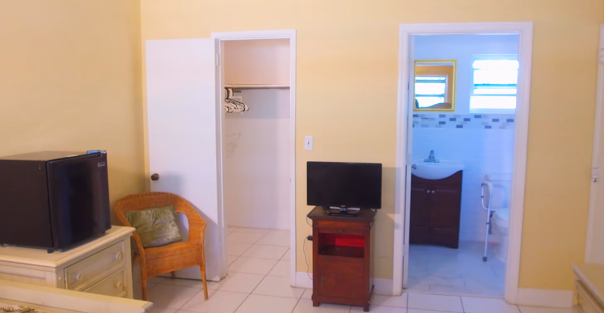 Interior view of a room with yellow walls featuring a small black refrigerator on a white dresser, a wicker chair with a green cushion, an open closet with hangers, a small wooden stand with a flat-screen TV, and a bathroom visible through an open door showing a sink and toilet with grab bars.
