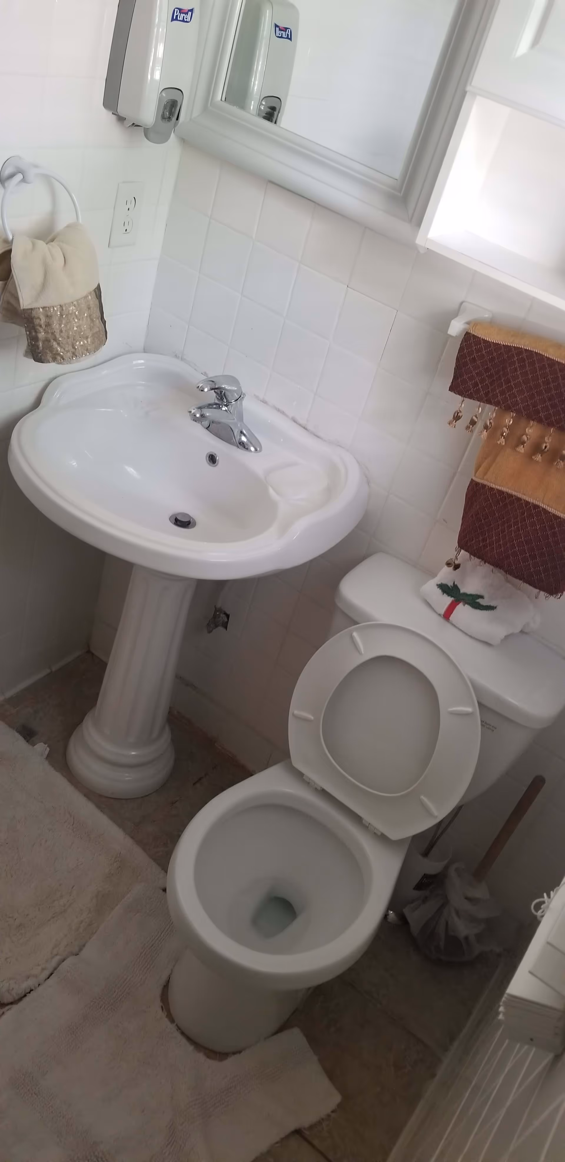 Pedestal sink and mirror next to an open toilet in a small tiled bathroom with towels and bath rugs.