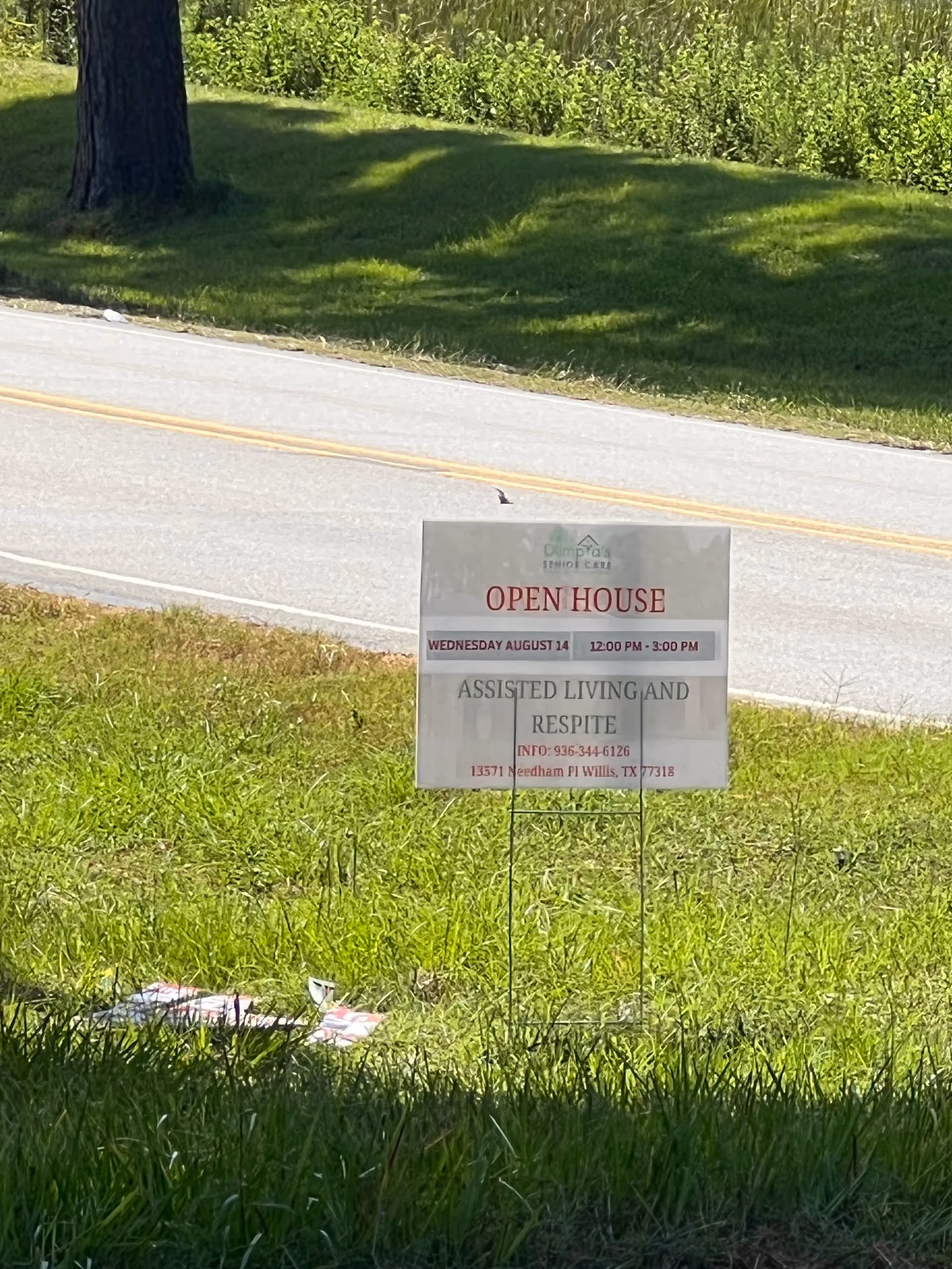 A lawn sign on a grassy area near a road advertising an open house event for assisted living and respite care at Forest Cottage Senior Care in Willis, Texas. The sign includes the date Wednesday August 14, the time 12:00 PM - 3:00 PM, a contact phone number, and the address 13571 Needham Pl Willis, TX 77318.