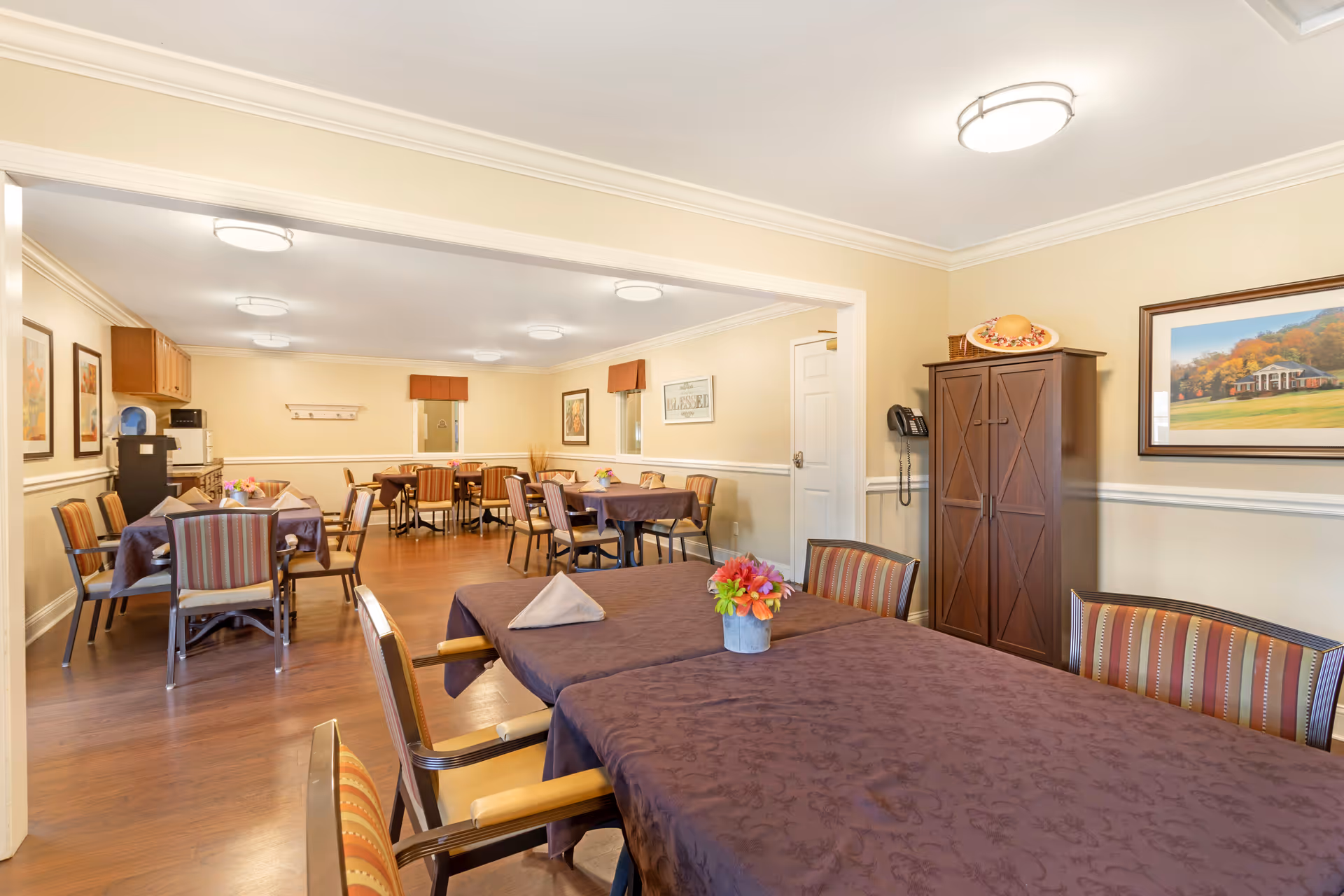 A dining room in a senior living facility with multiple tables covered in purple tablecloths, each set with napkins and small flower arrangements. The room has wooden floors, beige walls with white trim, framed artwork, and ceiling lights. There is a wooden cabinet and a wall-mounted telephone on the right side.