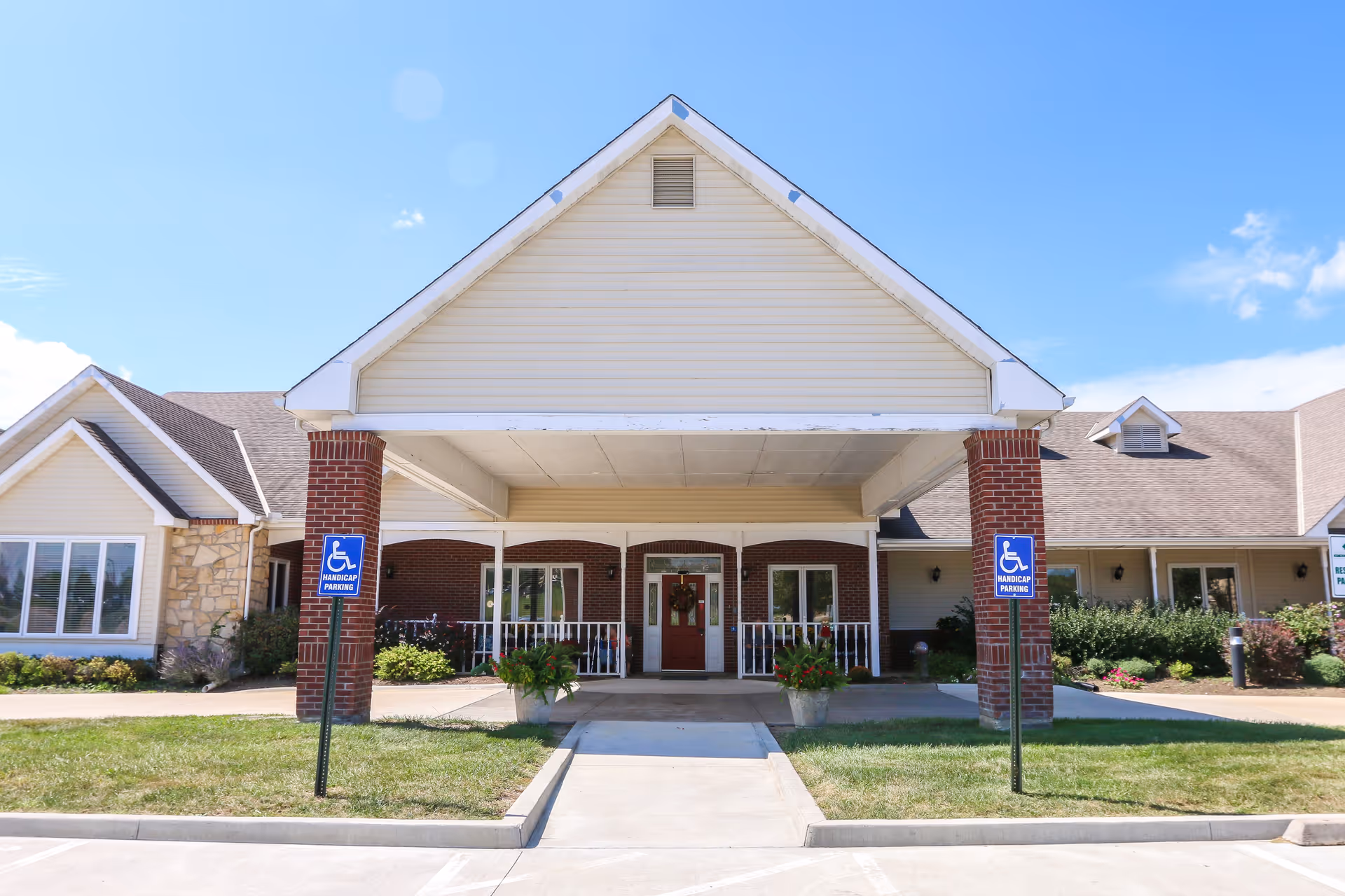 Front entrance of a single-story senior living building with a covered drive-through, brick columns, planters, and handicap parking signs.