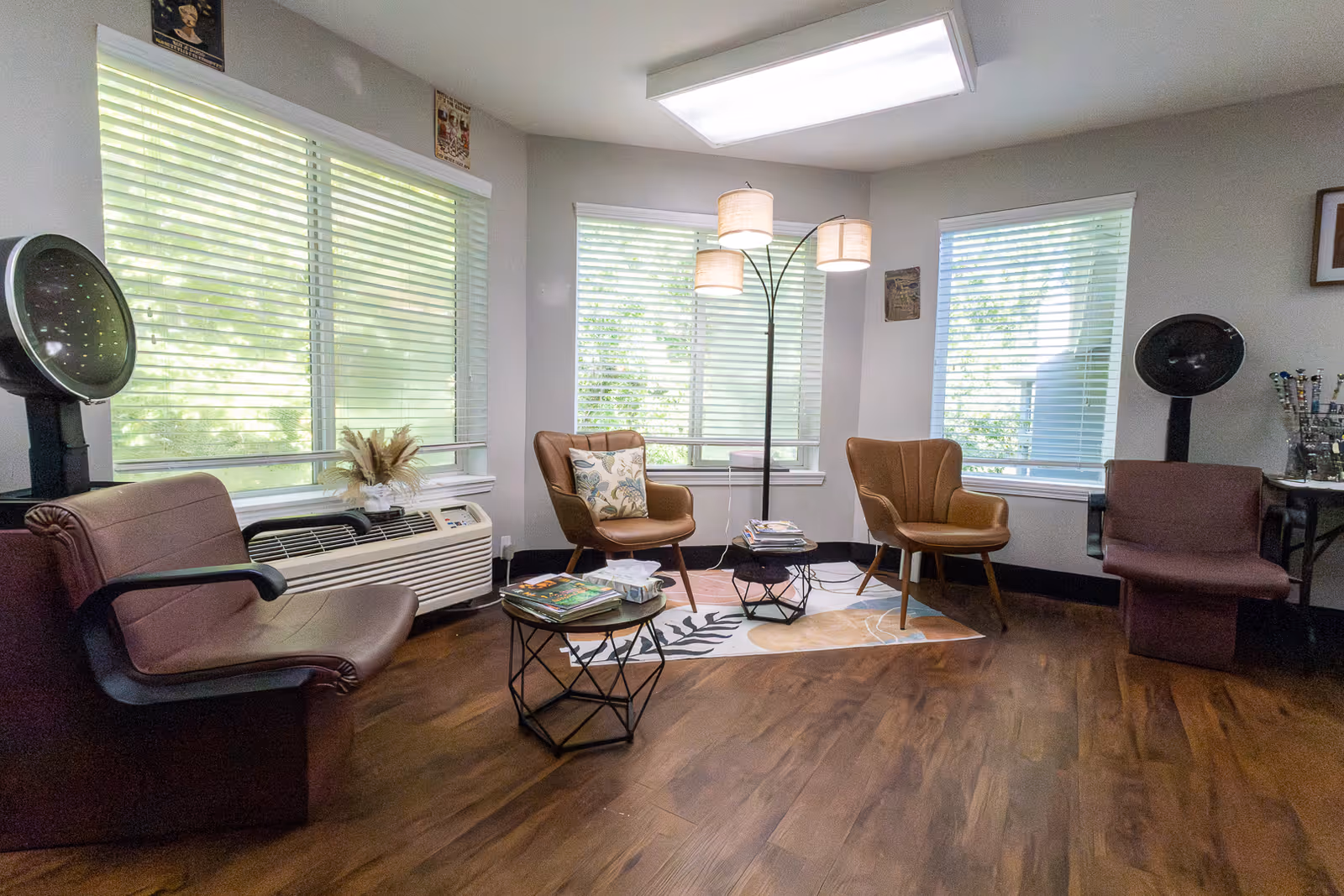 A cozy room with large windows covered by white blinds, featuring two brown leather chairs, two brown salon hair dryer chairs, a floor lamp with three lampshades, two small tables with magazines and tissues, and a wooden floor.